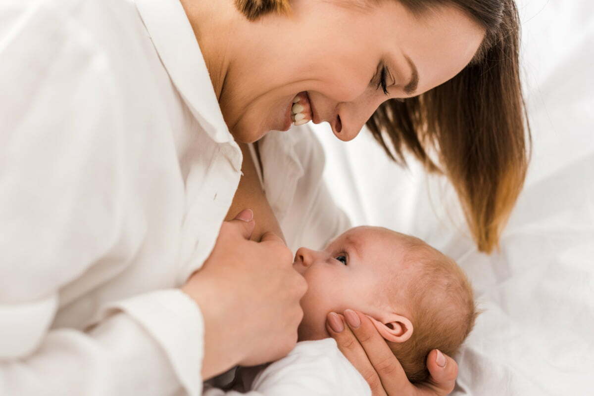 Smiling young mother in white t-shirt breastfeeding with implants
