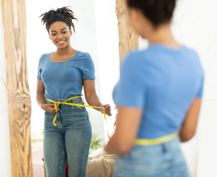 Happy Black Woman After Weight Loss Measuring Thin Waist With Tape Standing Near Mirror At Home.