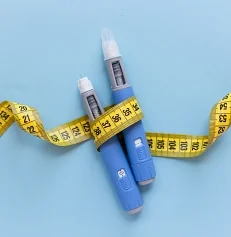 Two injectors dosing pens for subcutaneous injection of anti-obesity medication hovering over a blue background. Yellow measuring tape around the injectors.
