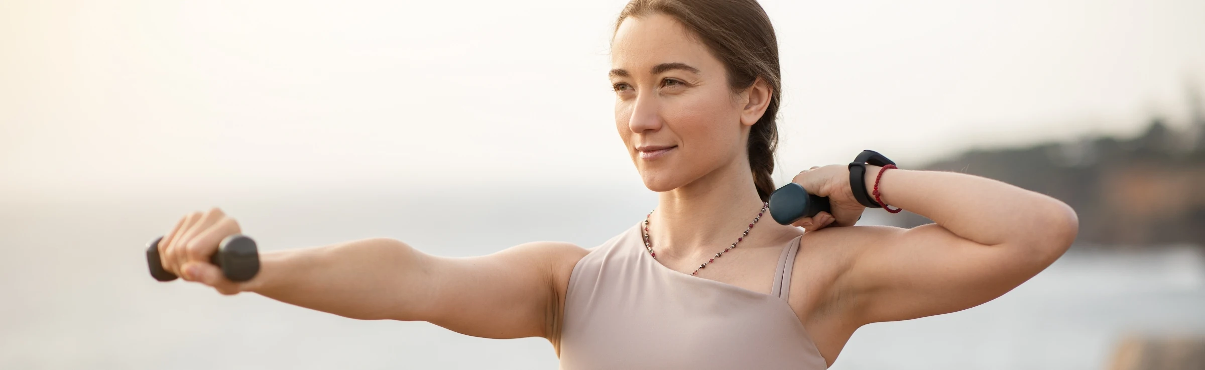Glad confident strong millennial caucasian woman athlete in sportswear doing arm exercises with dumbbells, enjoy workout, body care alone at sea beach.