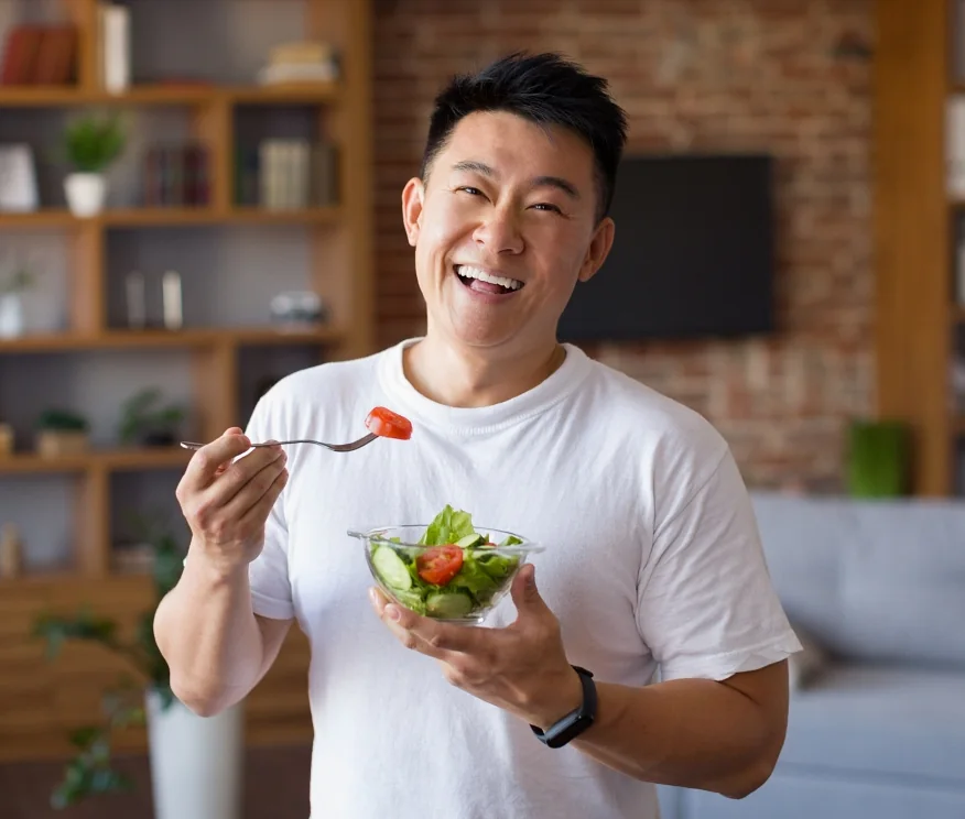 Happy asian mature man holding fork and bowl with fresh vegetable salad, eating healthy lunch after domestic training.